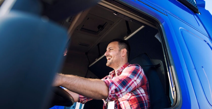 Professional middle aged trucker in cabin driving truck and smiling. Transportation industry.