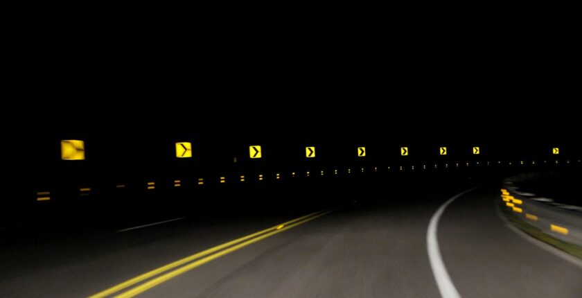 A series of yellow reflective 'sharp turn' signs being lit up by the lights of an oncoming car. The pitch black night sky is in contrast with the signs and street.